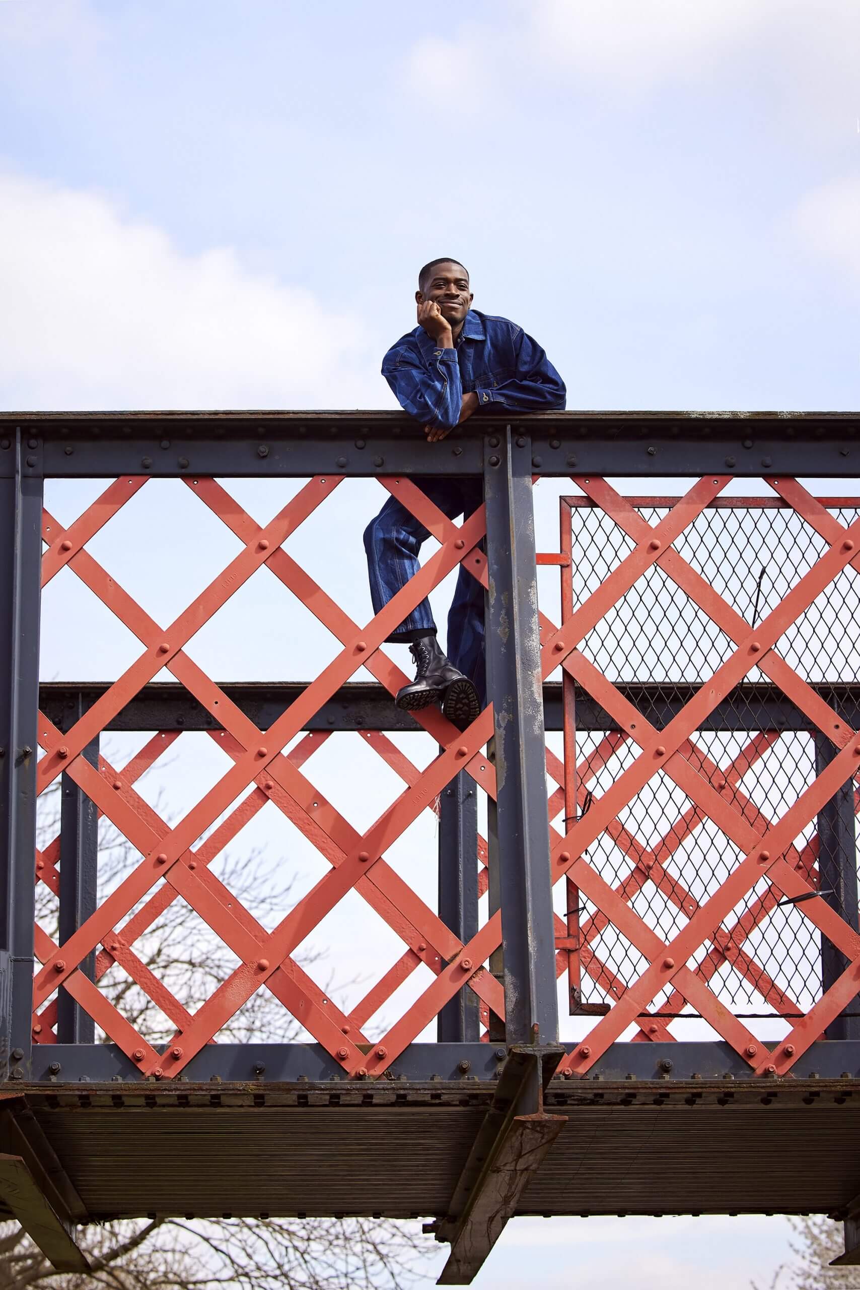 Damson Idris climbing up the metal railings on a bridge with his chin on his hand, smiling