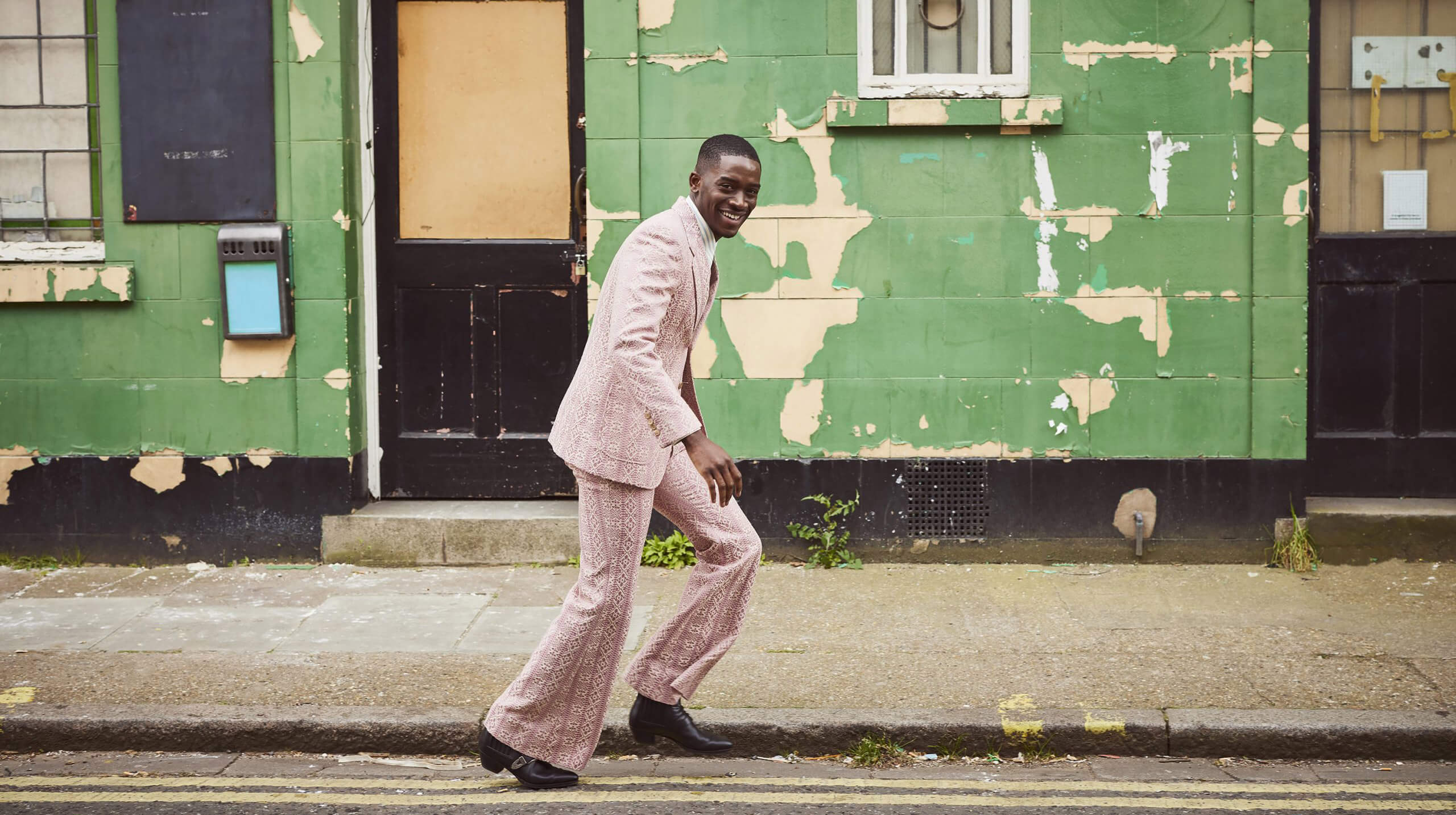 Damson Idris smiling while walking on the road by a flaking green painted wall