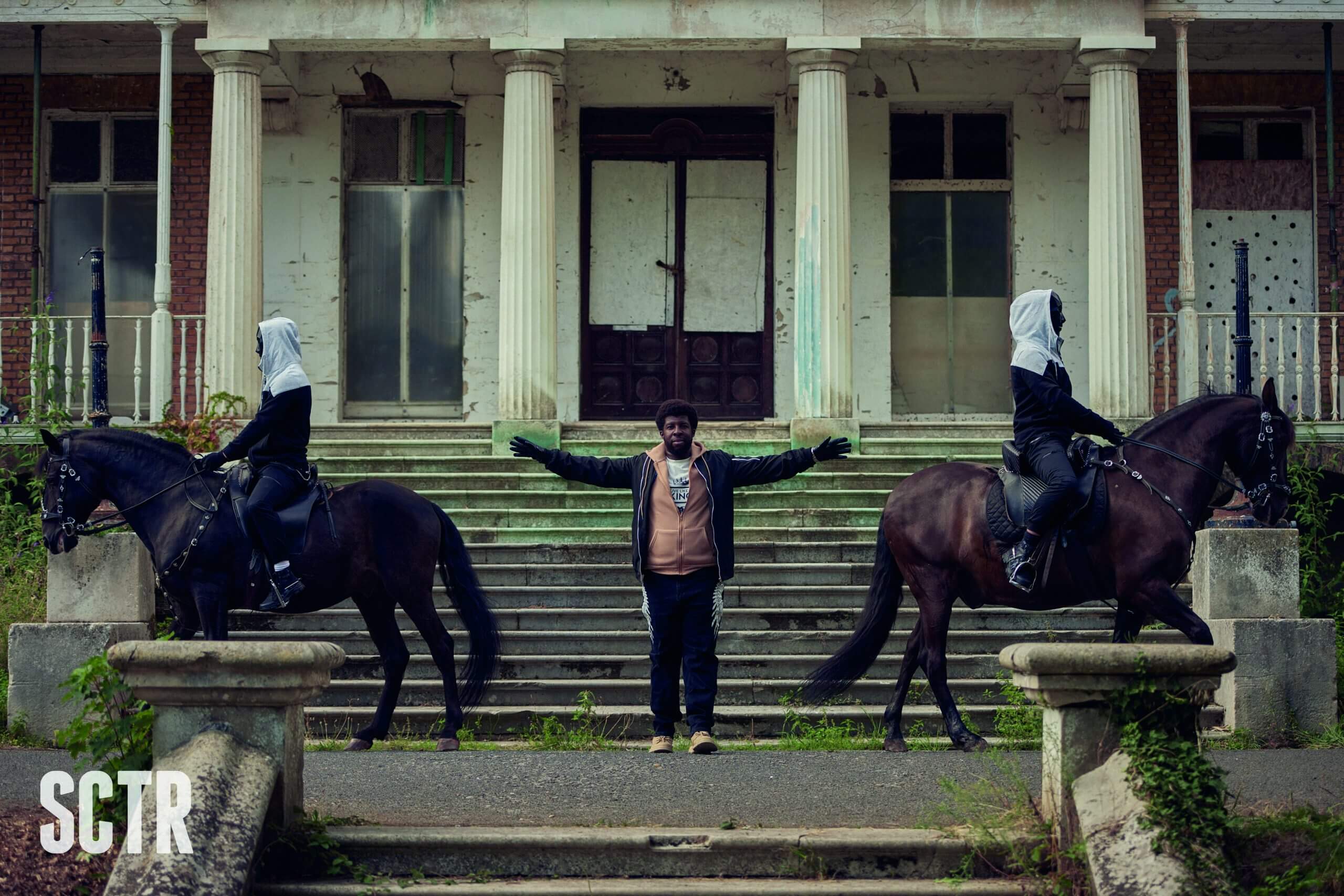 Aidie Palmer stood at the top of an outdoor staircase with arms out wide, two figures on horses are at his sides