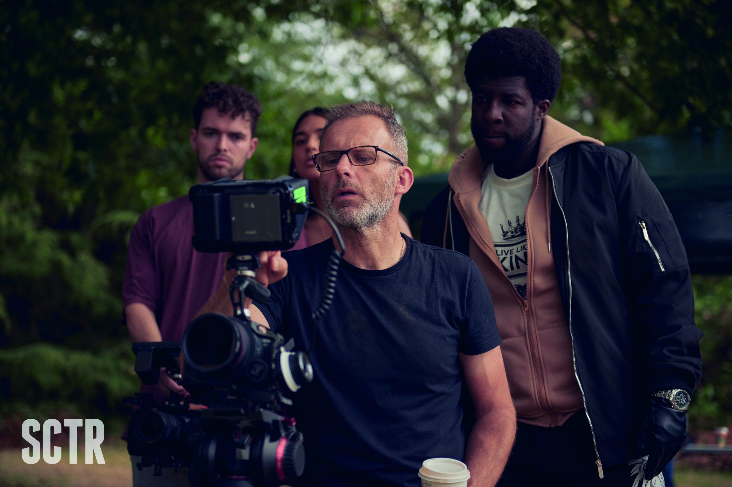 A camera operator studies a shot with 3 people stood behind him