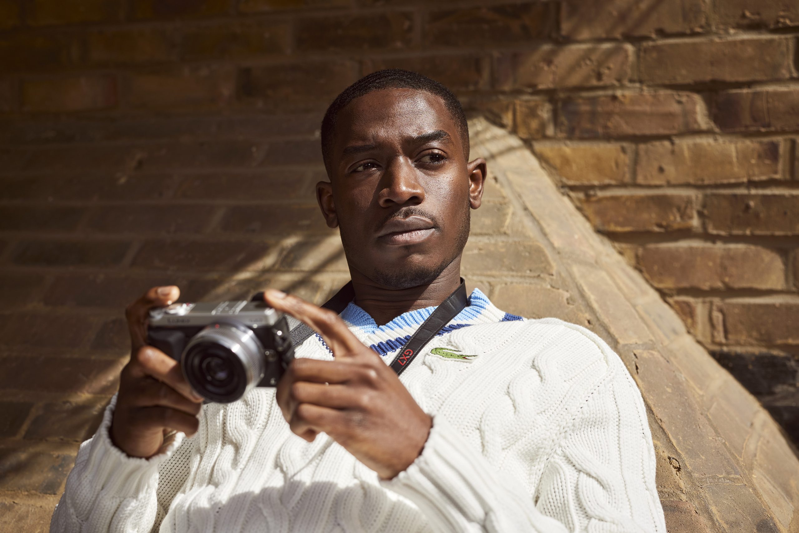 Damson Idris holding a camera while leaning on a brick wall