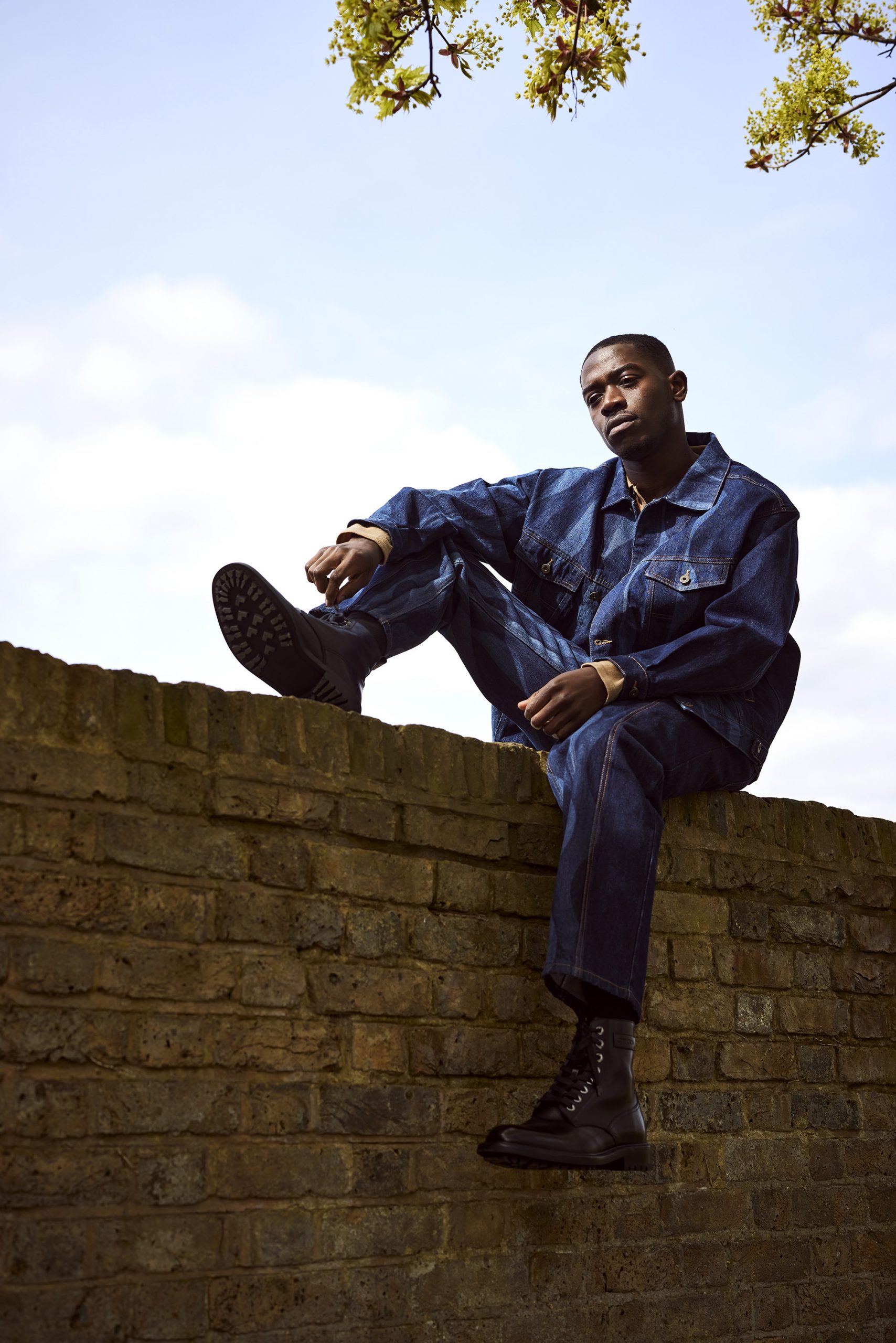 Damson Idris sat on a brick wall wearing a denim jacket and trousers